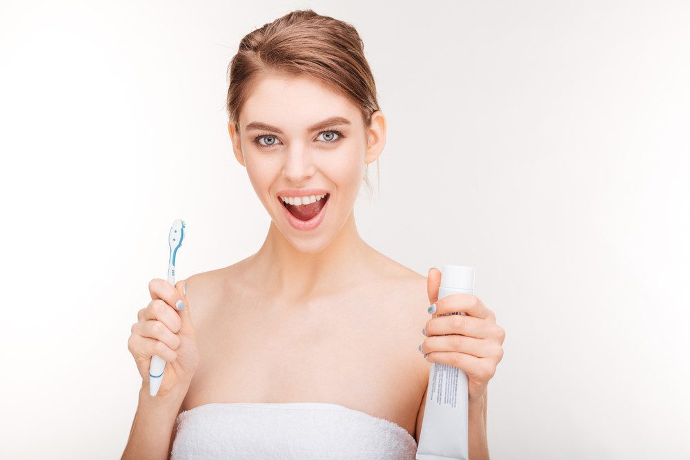 Beauty portrait of joyful charming young woman holding toothpaste and toothbrush over white background